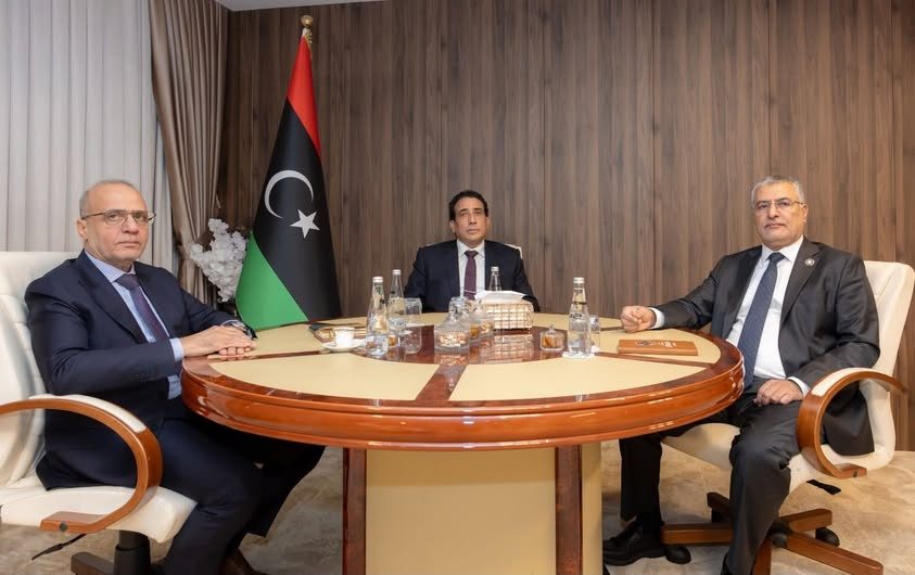 Three men seated at a round table in front of a Libyan flag