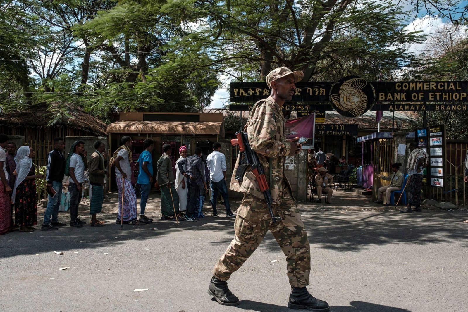 A soldier in camouflage walks along a street past people queuing outside a bank branch