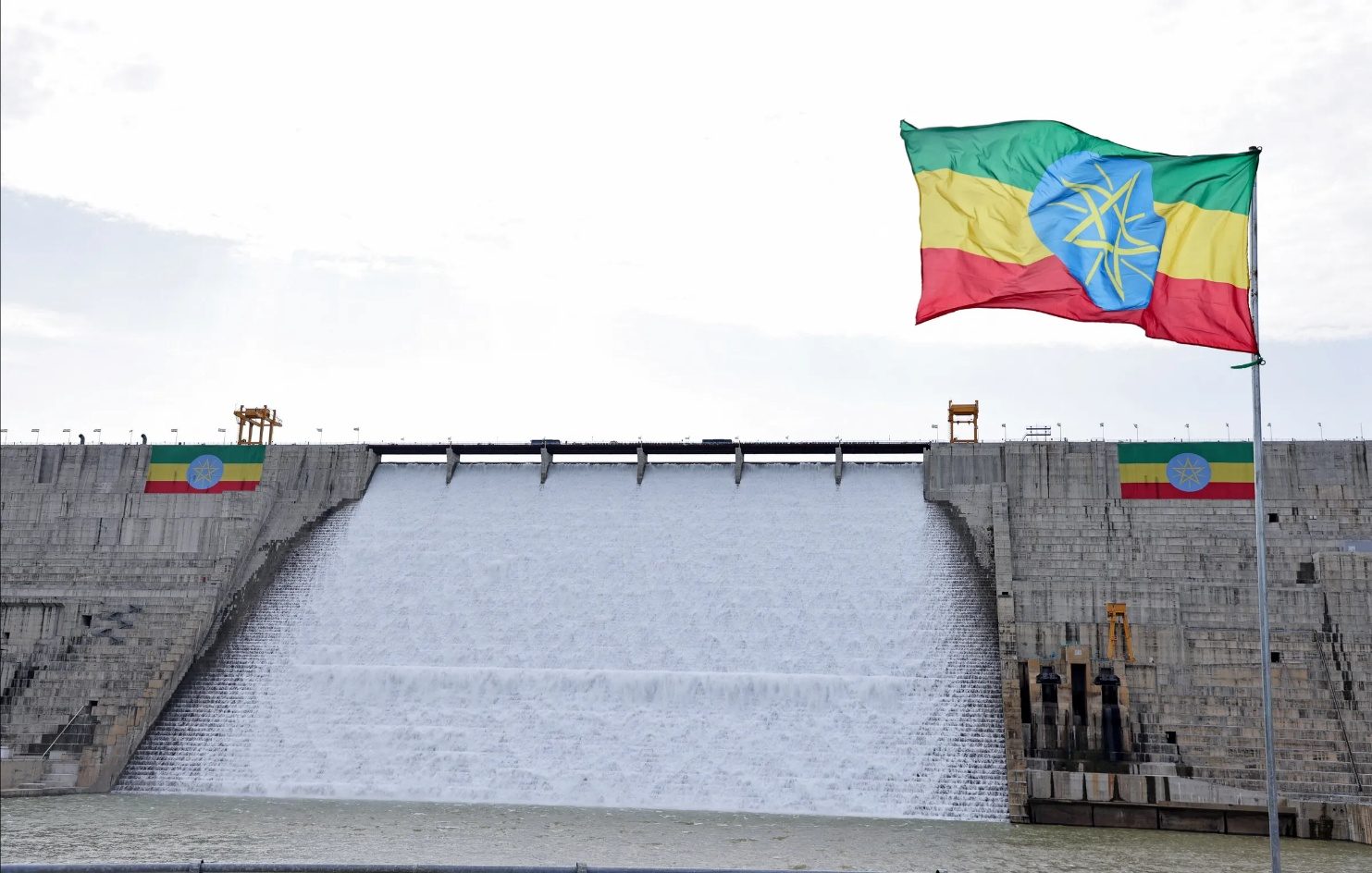Water pours over the spillway of a large dam with an Ethiopian flag flying beside it