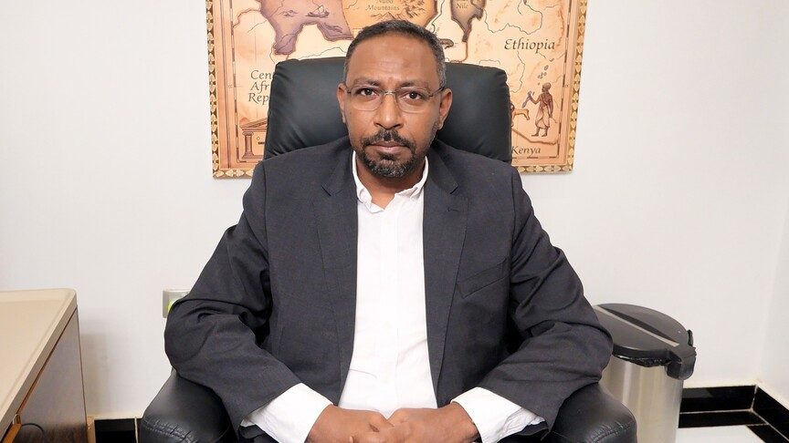 A man in a dark jacket and white shirt sits in an armchair in front of a wall map